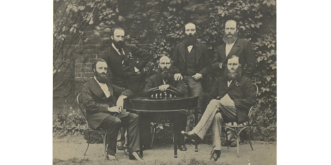 Black and white photo of six bearded men gathered around a table dressed in formal suits from the 1800s, threee seated and three standing