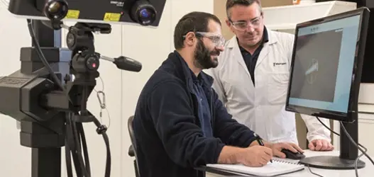 Two male scientists looking at white ceramic part on desktop screen