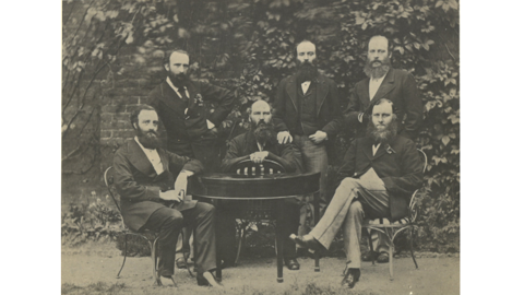 Black and white photo of six bearded men gathered around a table dressed in formal suits from the 1800s, threee seated and three standing